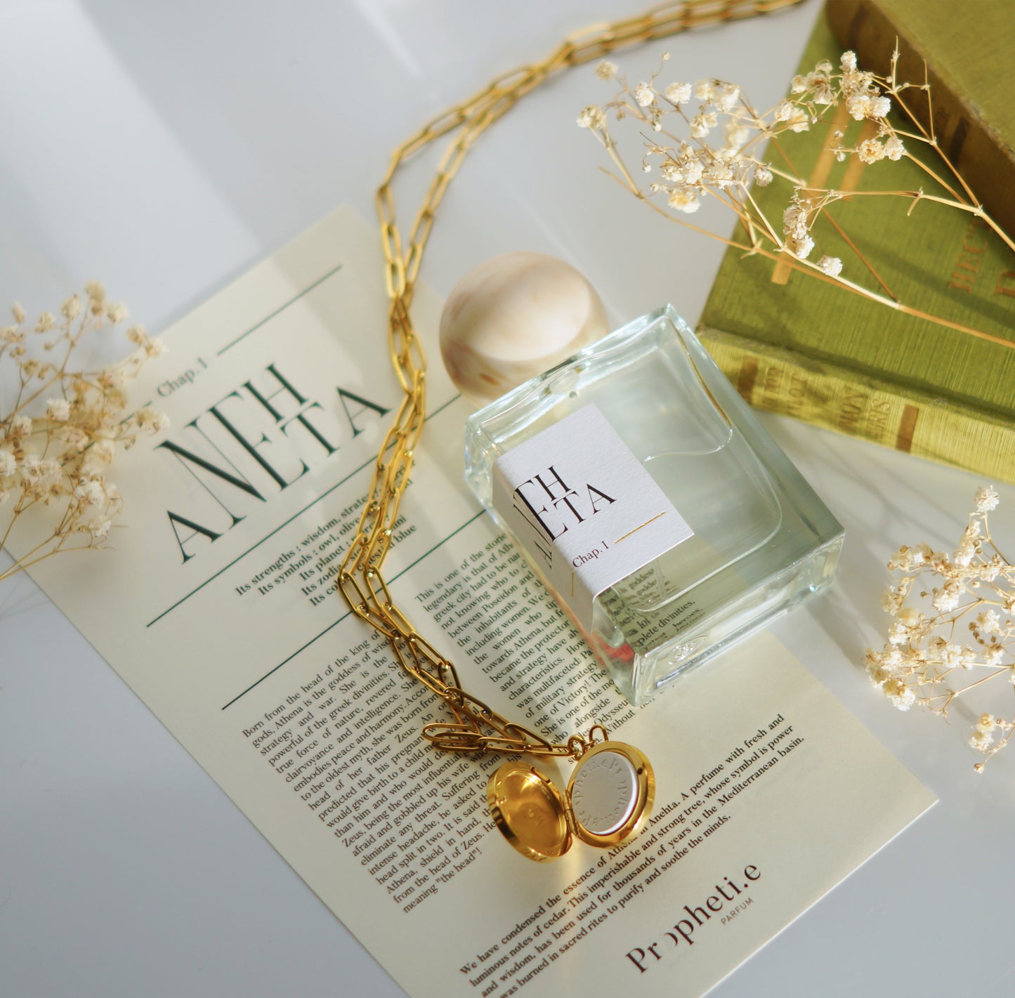 Parfum Anehta posé à plat sur le parchemin à plat, et le bijou posé dessus ouvert avec la pierre et quelques fleurs séchées ainsi que des livres